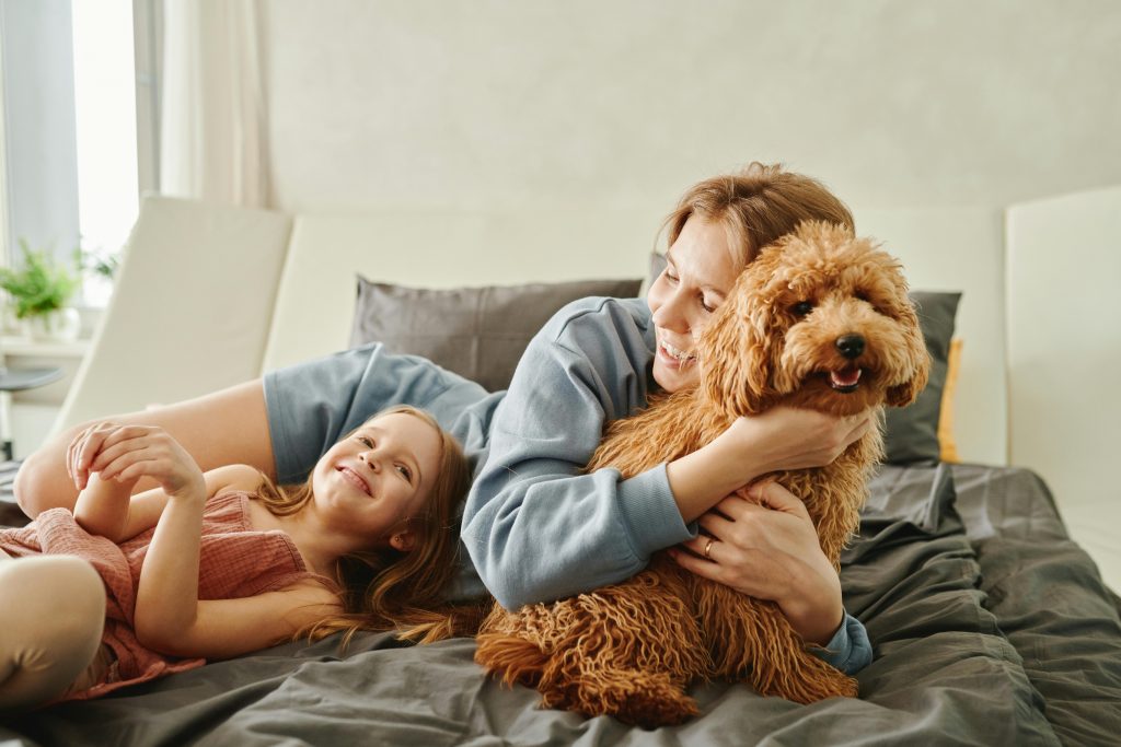 Frau und Kind genießen einen harmonischen Moment mit ihrem glücklichen Hund auf dem Bett – ein Bild, das das Wohlbefinden von Haustieren, die enge Bindung zwischen Tier und Halter*in und den Alltag voller Liebe und Fürsorge widerspiegelt.